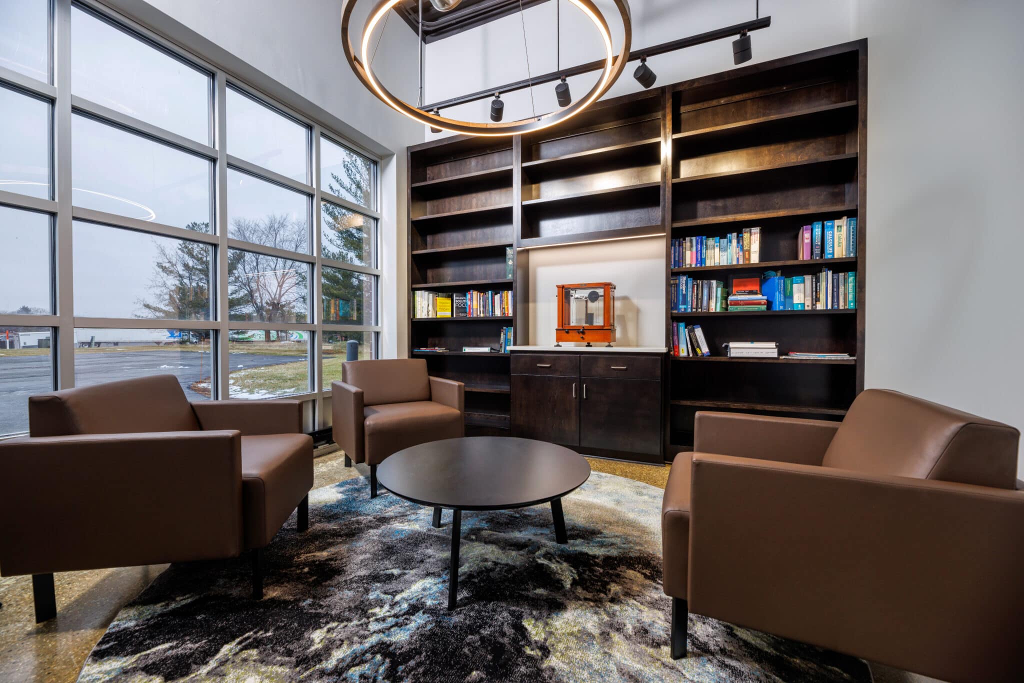 A modern interior at an office shows brown chairs, a wall-length bookshelf, and wall to ceiling windows to encourage natural light.